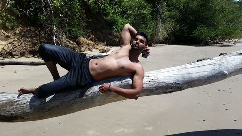 Portrait of shirtless man posing on tree trunk at beach