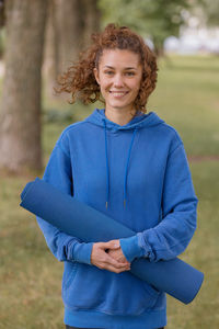 Portrait of smiling young woman holding exercise mat standing at park