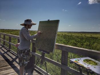 Side view of painter painting by landscape against sky