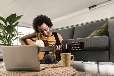 Smiling young woman at home with laptop playing guitar