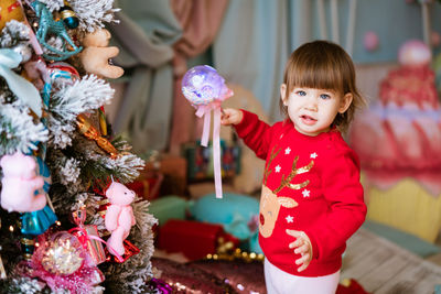 Portrait of little girl in red christmas sweater stands by christmas tree