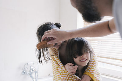 Man combing hair of daughter in towel with sister in bathroom