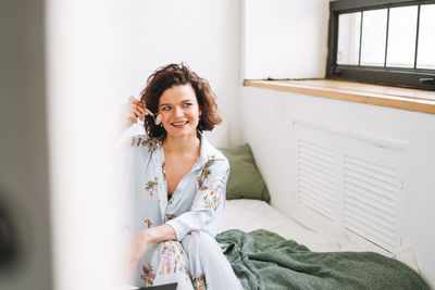 Young brunette woman in blue pajamas doing face massage with stone roller sitting on bed at home