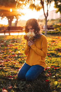 Curly young girl in yellow sweater on grass smells autumn bouquet of dry leaves and flowers
