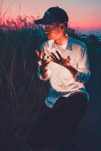 Young man with illuminated string light by grass against sky during sunset