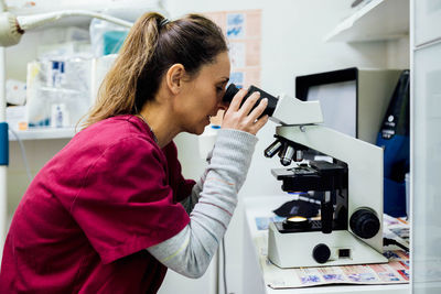 Side view of focused female veterinarian in uniform looking through microscope while checking results of medical procedure in modern clinic