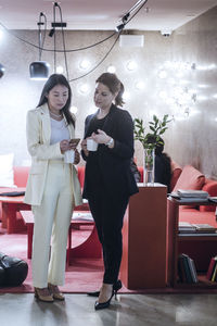 Full length of female coworkers talking during coffee break while colleagues working in background