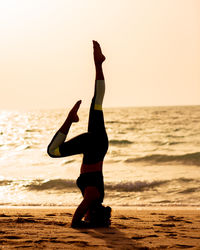 Full length of woman practicing headstand at beach against clear sky during sunset