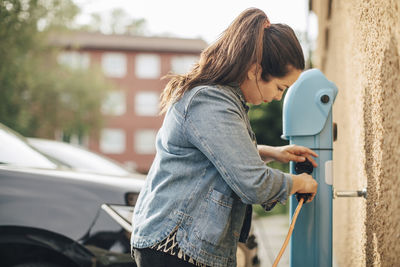 Side view of woman adjusting electric plug for charging car at station