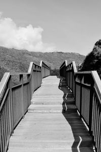 Empty wooden footbridge leading towards water against sky