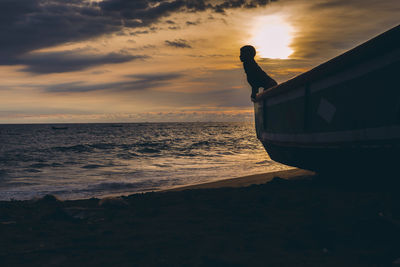 Silhouette boy in boat at beach against sky during sunset