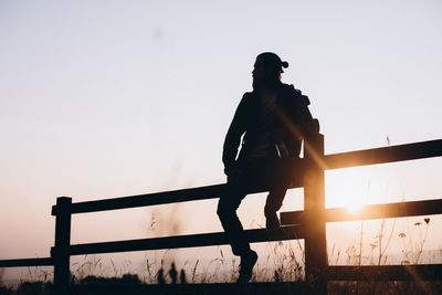 Silhouette man sitting on railing against clear sky during sunrise