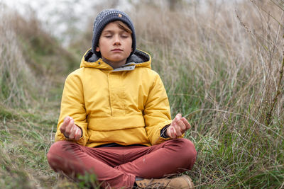 Small boy doing yoga relaxation exercises in lotus position during autumn day in tall grass.
