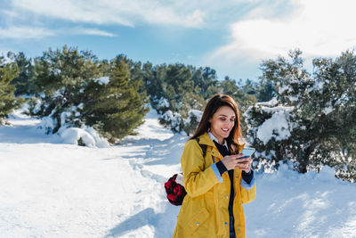 Young woman using phone while standing in snow against sky during winter