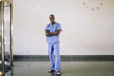 Portrait of smiling male doctor standing with arms crossed in front of white wall at hospital