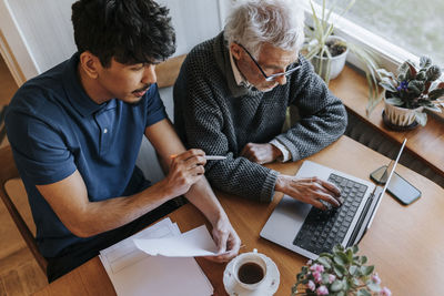 Senior man using laptop while sitting next to male caregiver explaining medical reports at home