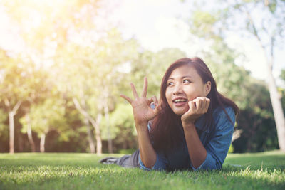 Happy young woman showing ok sign while lying on grass at park