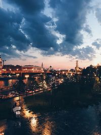 Illuminated buildings by river against sky at sunset