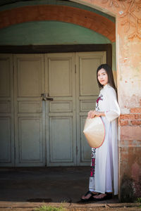 Portrait of smiling young woman holding hat against building