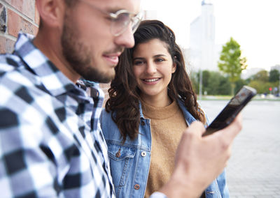 Smiling woman looking at friend using mobile phone against wall