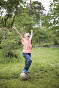 Excited boy shouting with arms raised while standing on soccer ball at back yard