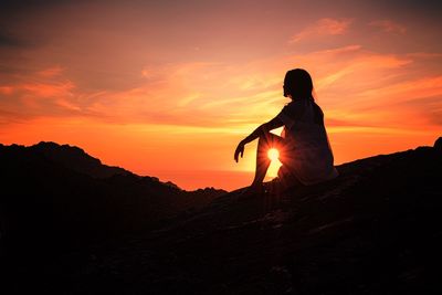 Silhouette woman sitting on mountain against orange sky