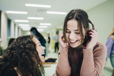 Cheerful teenage girl with headphones sitting with female classmate in corridor at high school