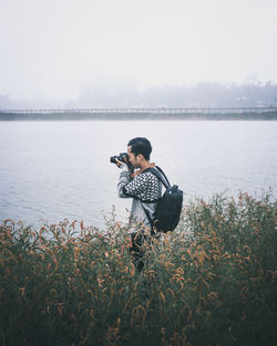 Side view of man photographing with digital camera while standing amidst plants by lake