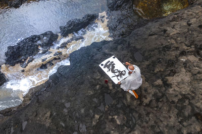 Mature monk doing calligraphy sitting on rock by sea