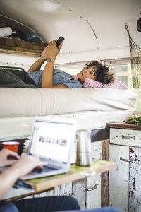 Young woman lying on bed while using mobile phone by man with laptop in caravan