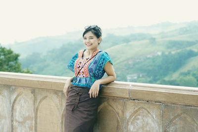 Portrait of smiling young woman leaning on railing against mountains