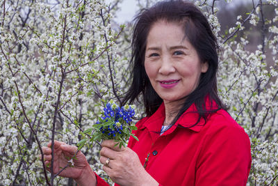 Portrait of smiling woman holding red flowering plant against trees