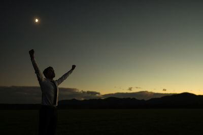 Man with arms raised standing on land against sky during sunset