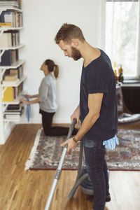 Side view of man vacuuming floor while woman cleaning shelves in background at home