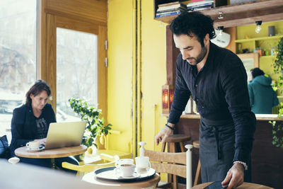 Waiter working in cafe with customer using laptop in background