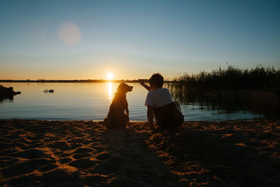 Friends sitting on shore at beach against sky during sunset