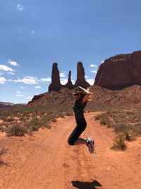 Woman jumping over dirt road against blue sky