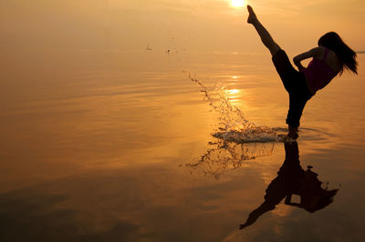 Silhouette of woman kicking while standing in sea against sky during sunset