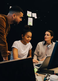 Young multi-ethnic business colleagues discussing over laptop against bulletin board at office