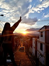 Silhouette woman with arms raised standing in city against sky during sunset
