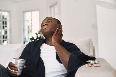 Tired man looking away while sitting with coffee cup on sofa at home