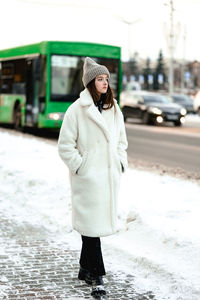 A young girl in a white fur coat in winter stands at a bus stop and waits for her bus