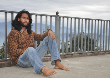 A young man with long hair and beard looking at camera while sitting with leaning on safety barrier 