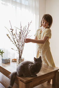 A happy girl hangs easter eggs on a willow bouquet at home with a little kitten. funny pets. easter