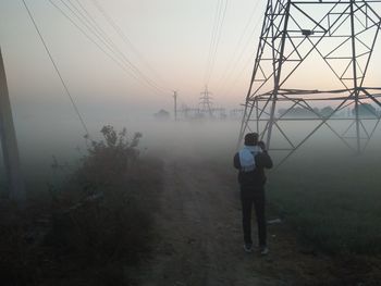 Rear view of man standing on electricity pylon in foggy weather