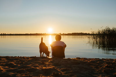 Rear view of men sitting on beach against sky during sunset