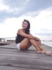 Portrait of smiling young woman sitting on pier against sky