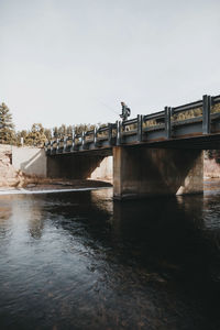 Low angle view of hiker with fishing rod standing on bridge over river against clear sky