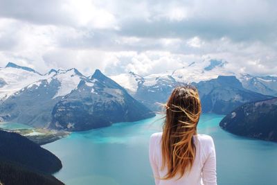 Rear view of woman looking at lake by snowcapped mountains against cloudy sky