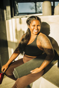 Happy woman with exercise mat sitting near wall after yoga class